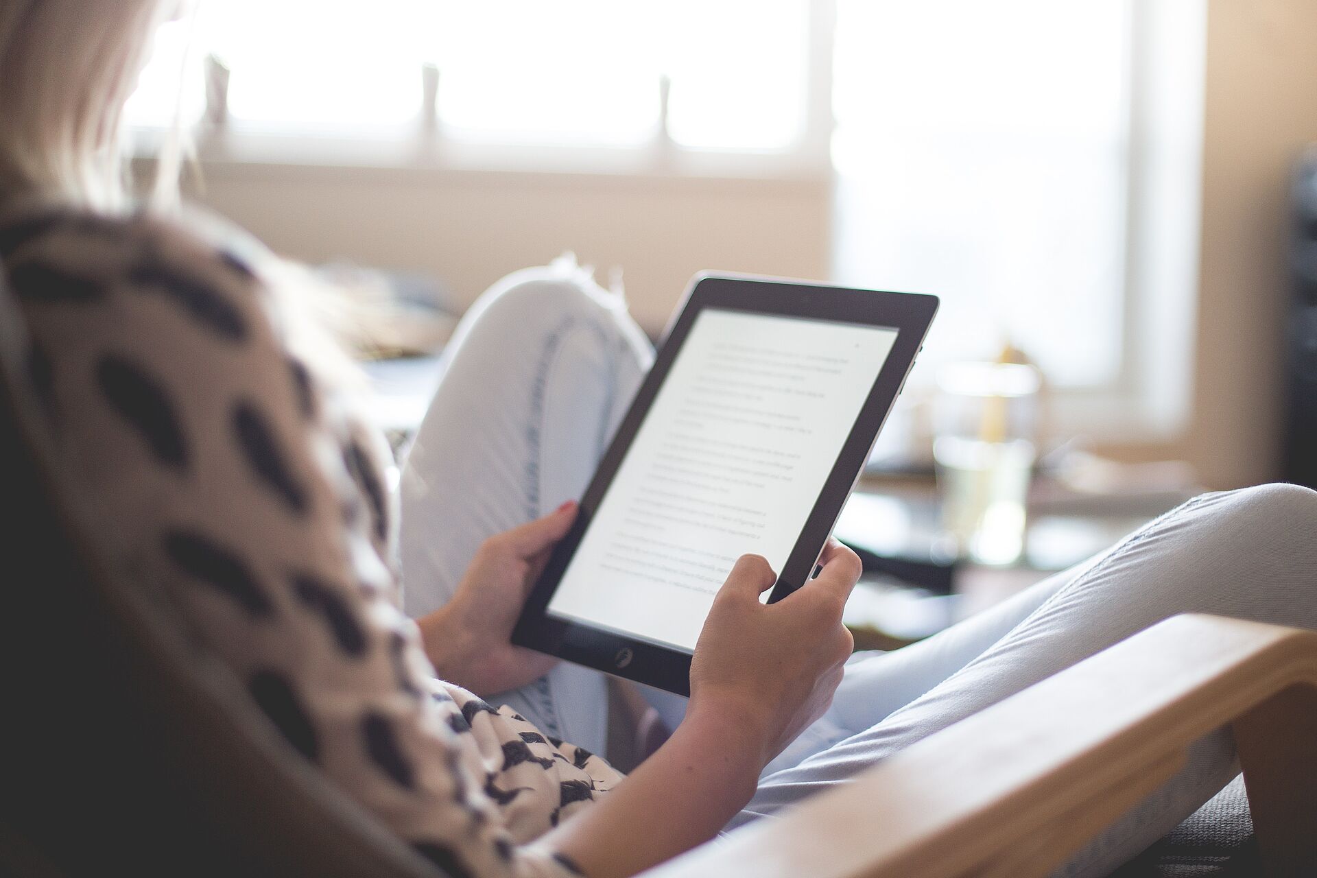 Person holding a tablet and reading a text, sitting relaxed in a bright living room or reading environment with a blurred background.