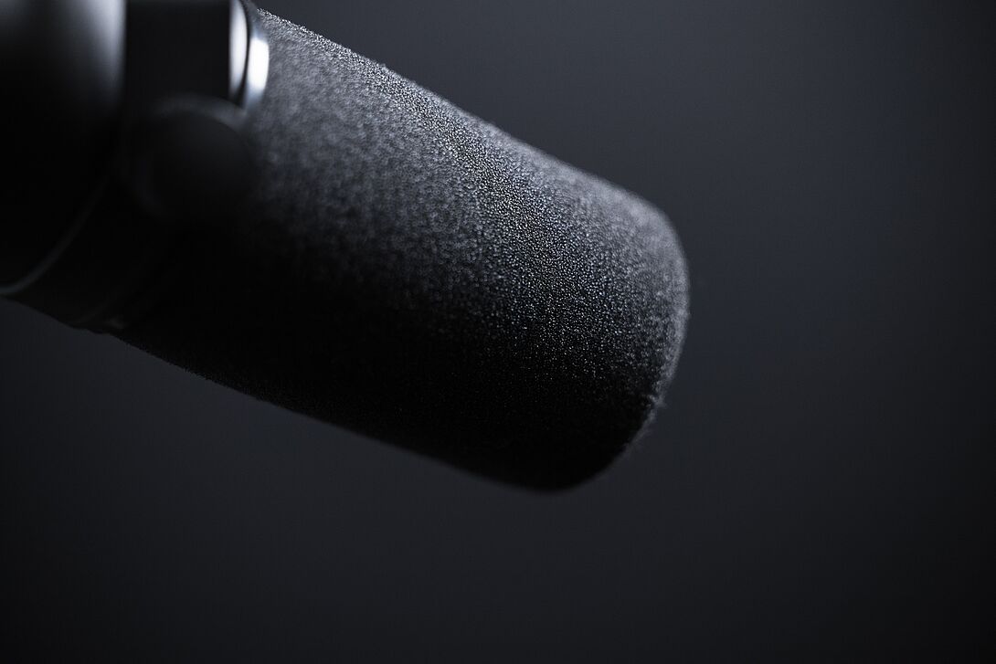 Close-up of a black studio or podcast microphone with foam windscreen against a dark, neutral background.